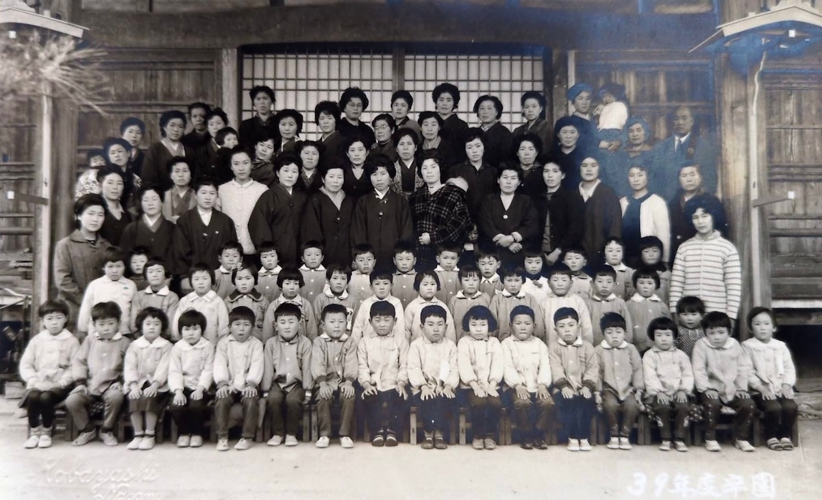 A group photo from the kindergarten graduation ceremony. Wakatsuki's note states that there was a mumps epidemic at the time, and everyone looked big-headed. Wakatsuki is pictured in front of the woman with the borders on the far right.